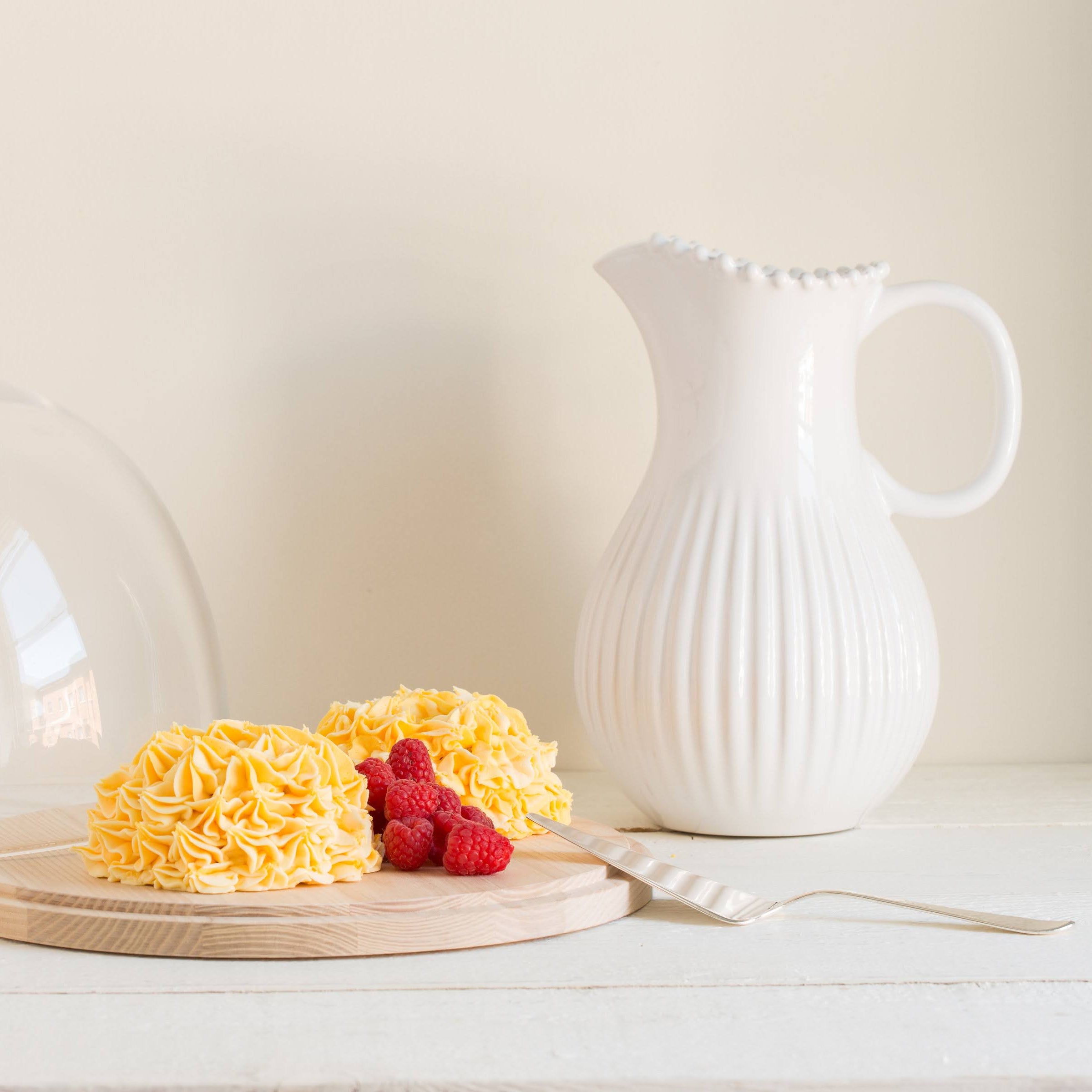 Yellow cake with raspberries under a glass cloche on a wooden board, next to a white ceramic pitcher.