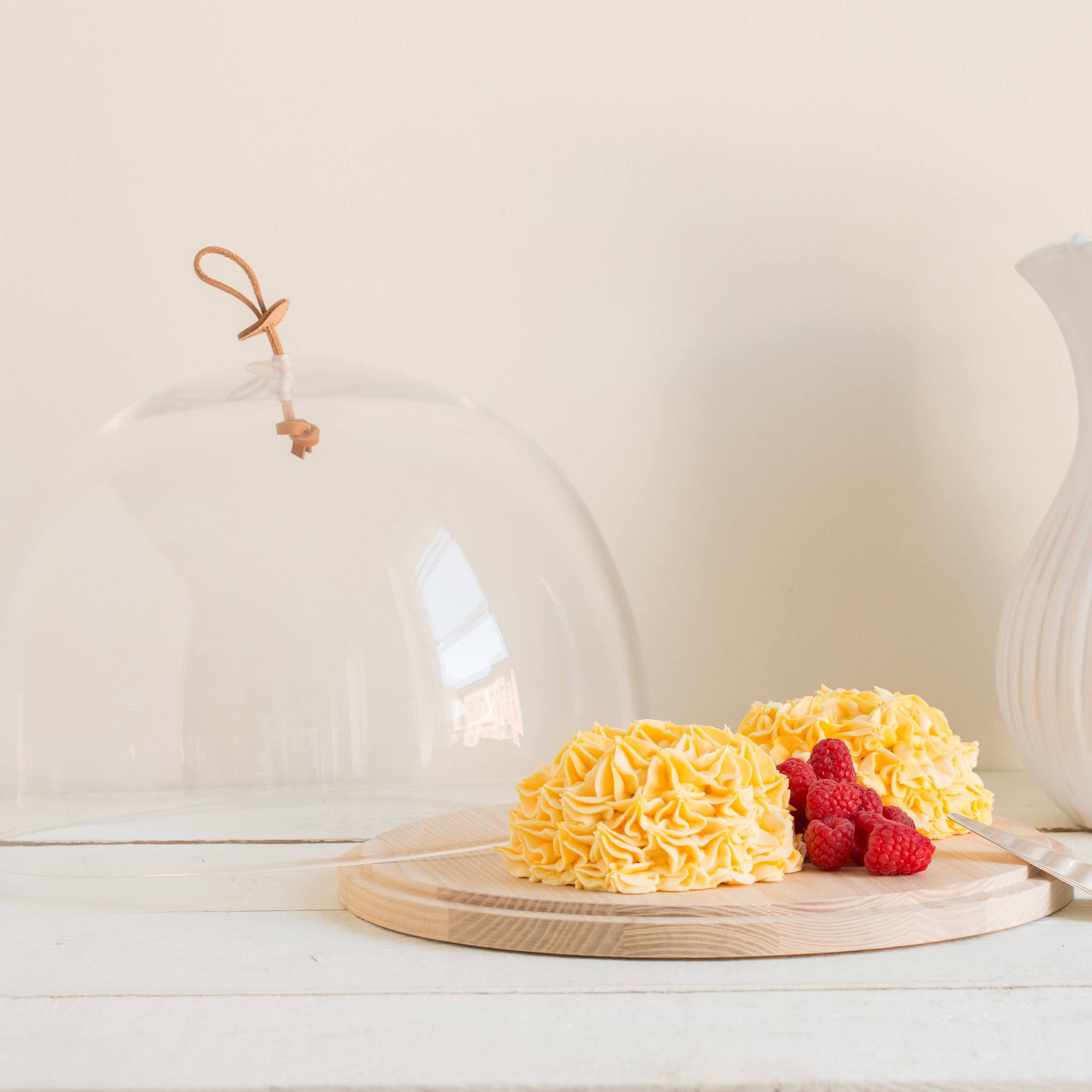 Yellow cake with raspberries under a glass cloche on a wooden board, next to a white ceramic pitcher.