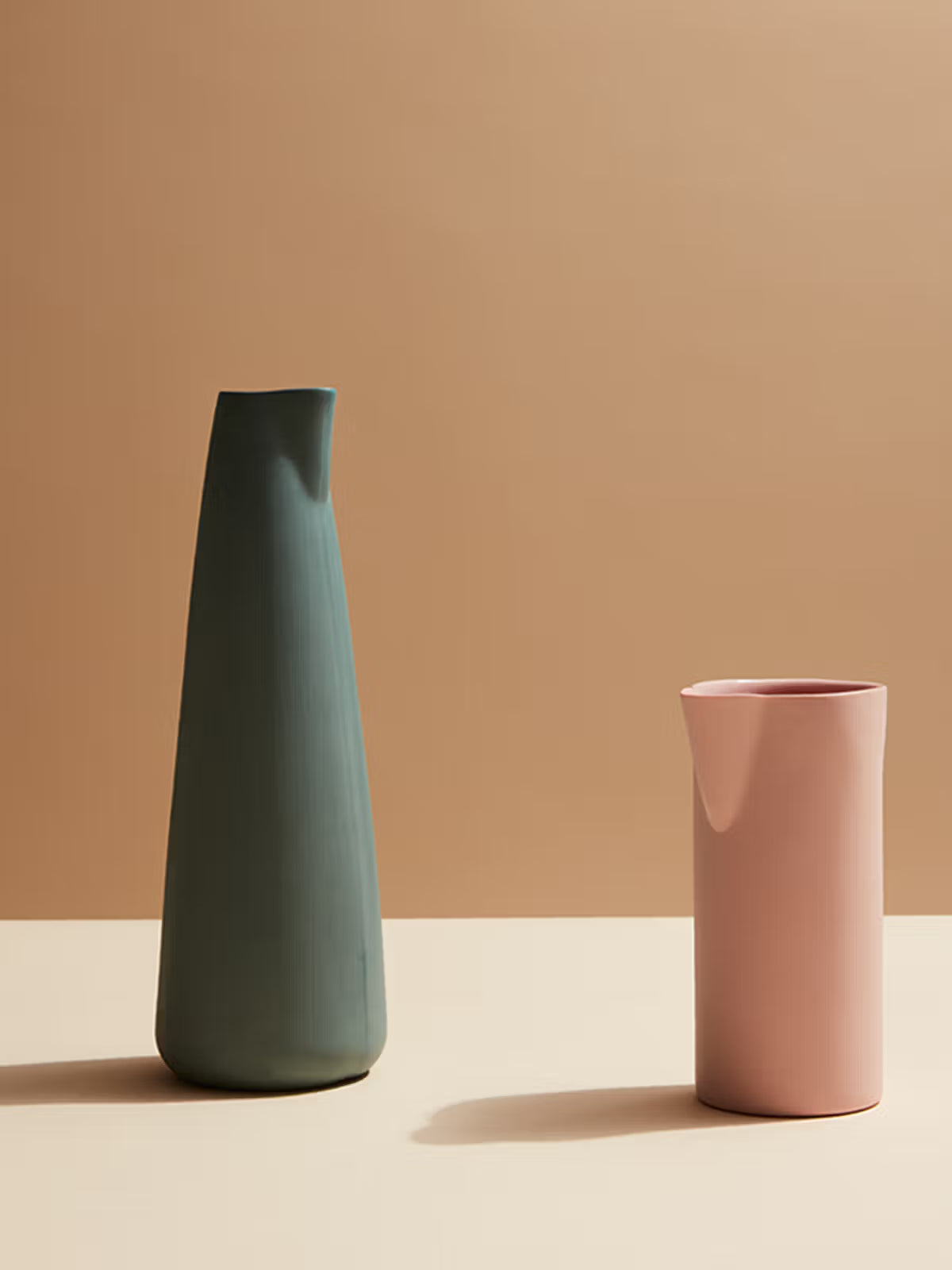 Two ceramic vases, one green and one pink, on a beige surface with a brown background.