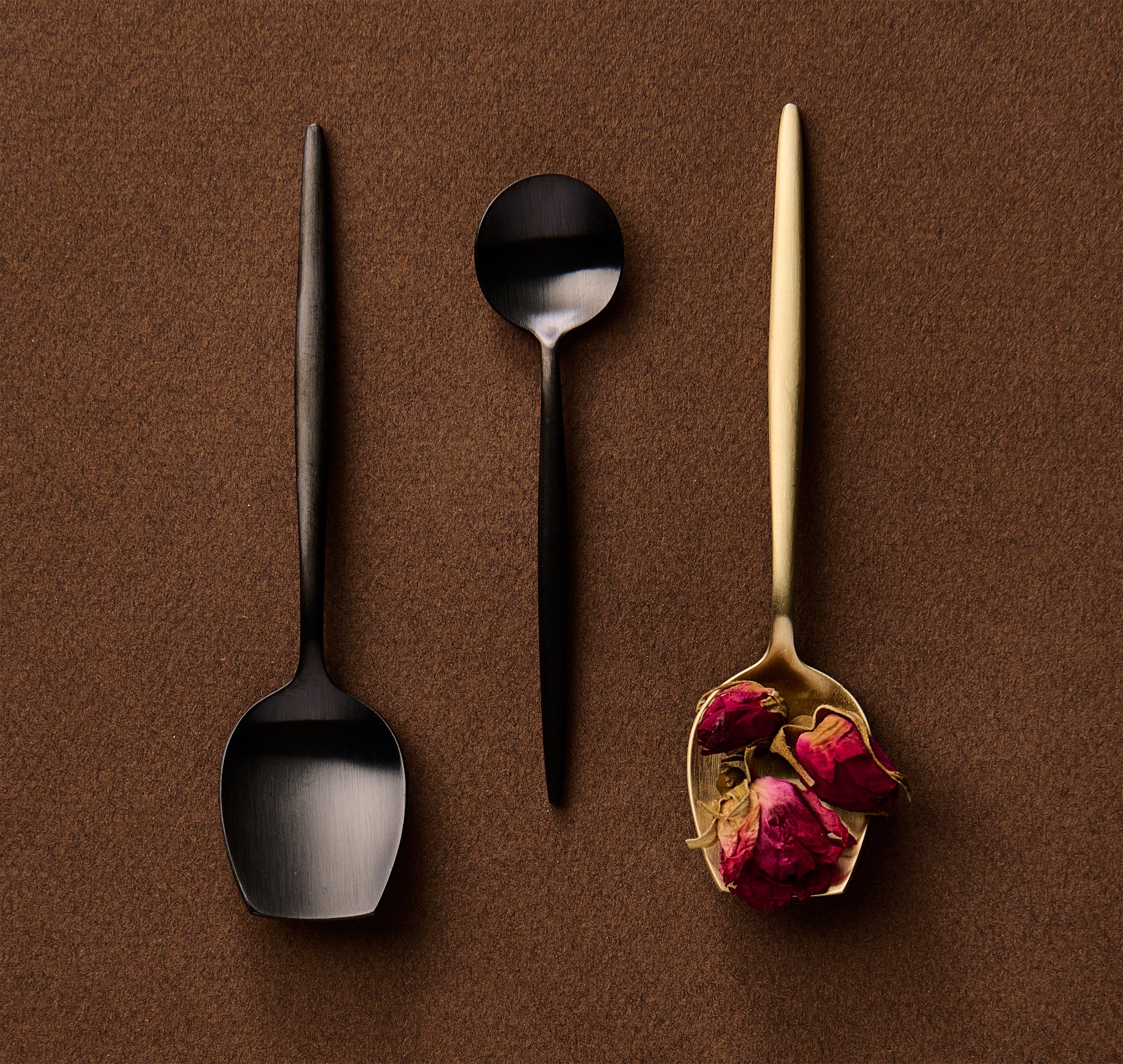 Three spoons on a brown background, two black and one gold with dried flowers.
