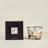Decorative candle with a scenic design next to its packaging on a light beige background