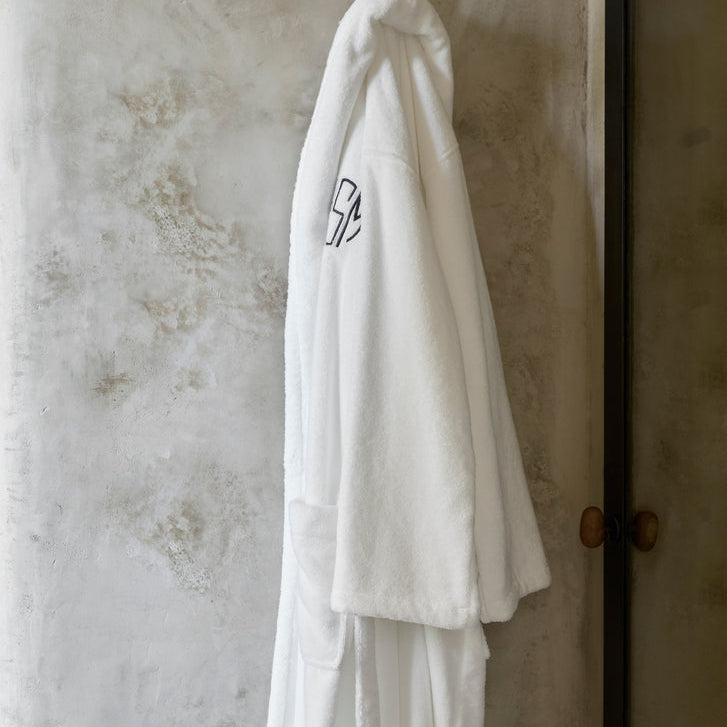 White bathrobe with a logo hanging on a hook against a textured wall.