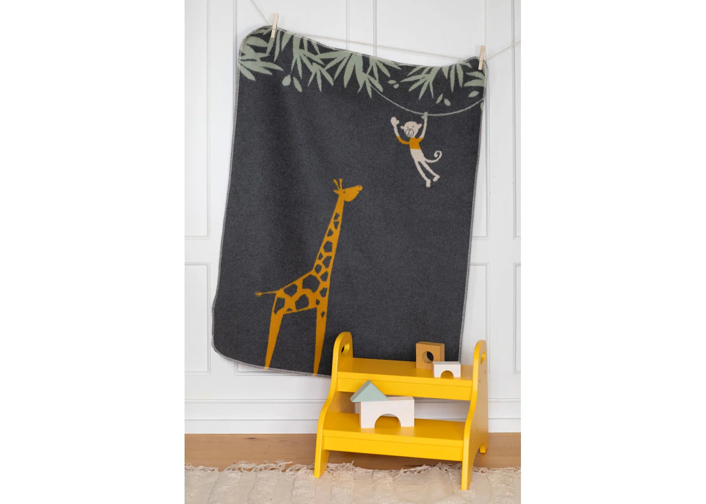 Children's room with a dark gray wall hanging featuring a giraffe and monkey, a yellow shelf with toys, and a white wall.
