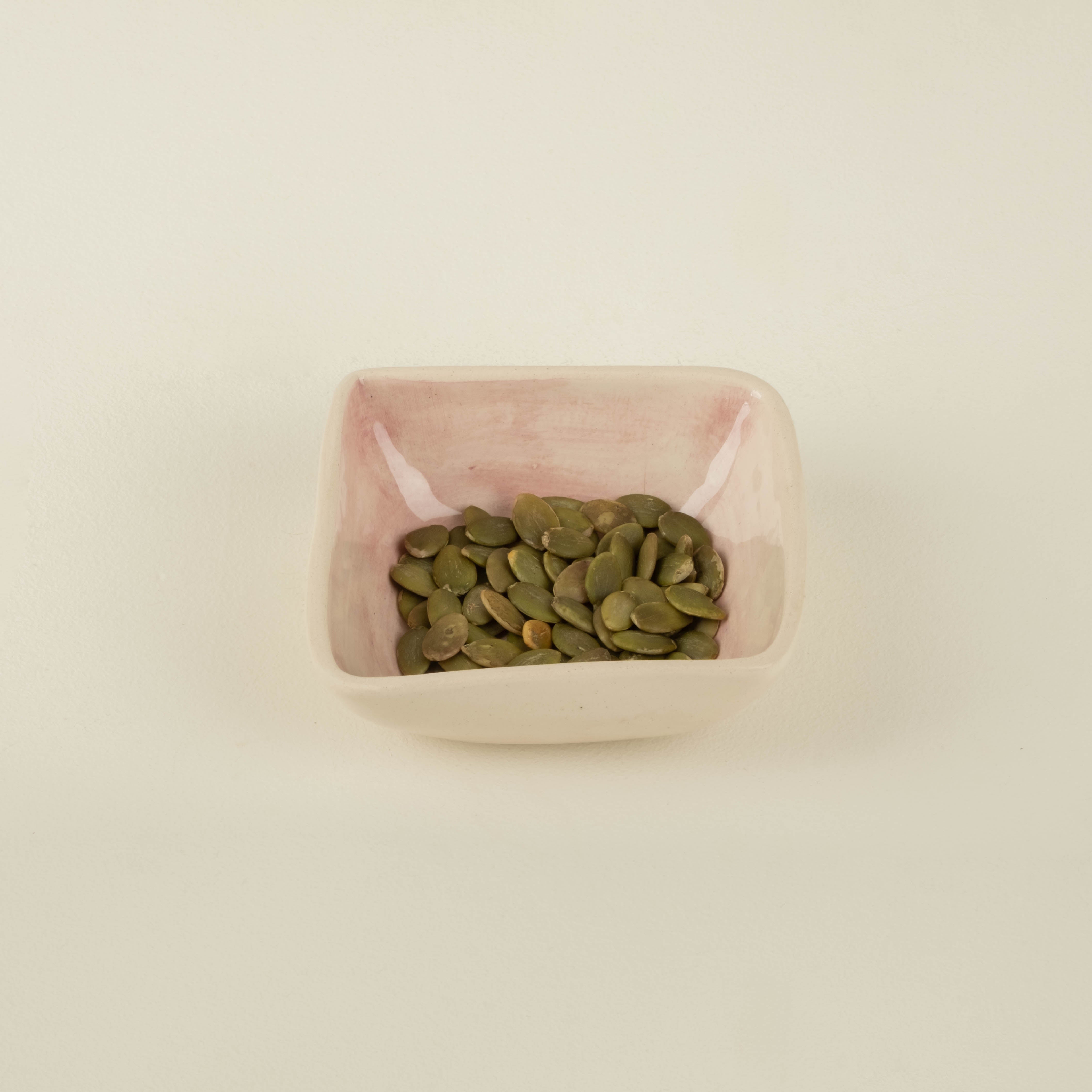 Small pink bowl filled with green pumpkin seeds on a beige background
