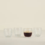 Four glass cups on a beige background, with one containing a dark liquid.