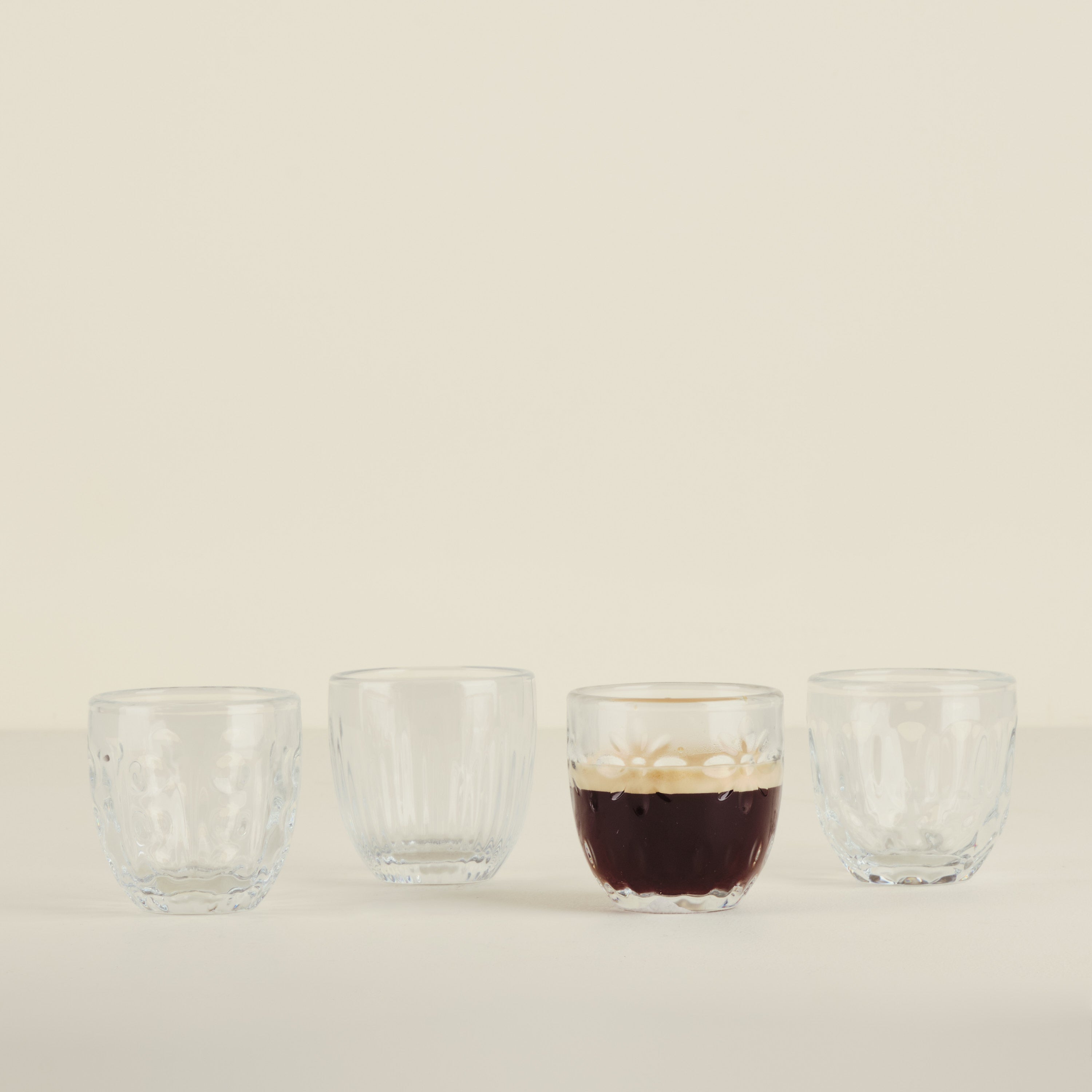 Four glass cups on a beige background, with one containing a dark liquid.