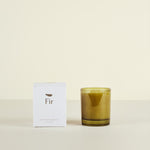 Green candle in a glass holder with a white box labeled 'Fir' on a beige background