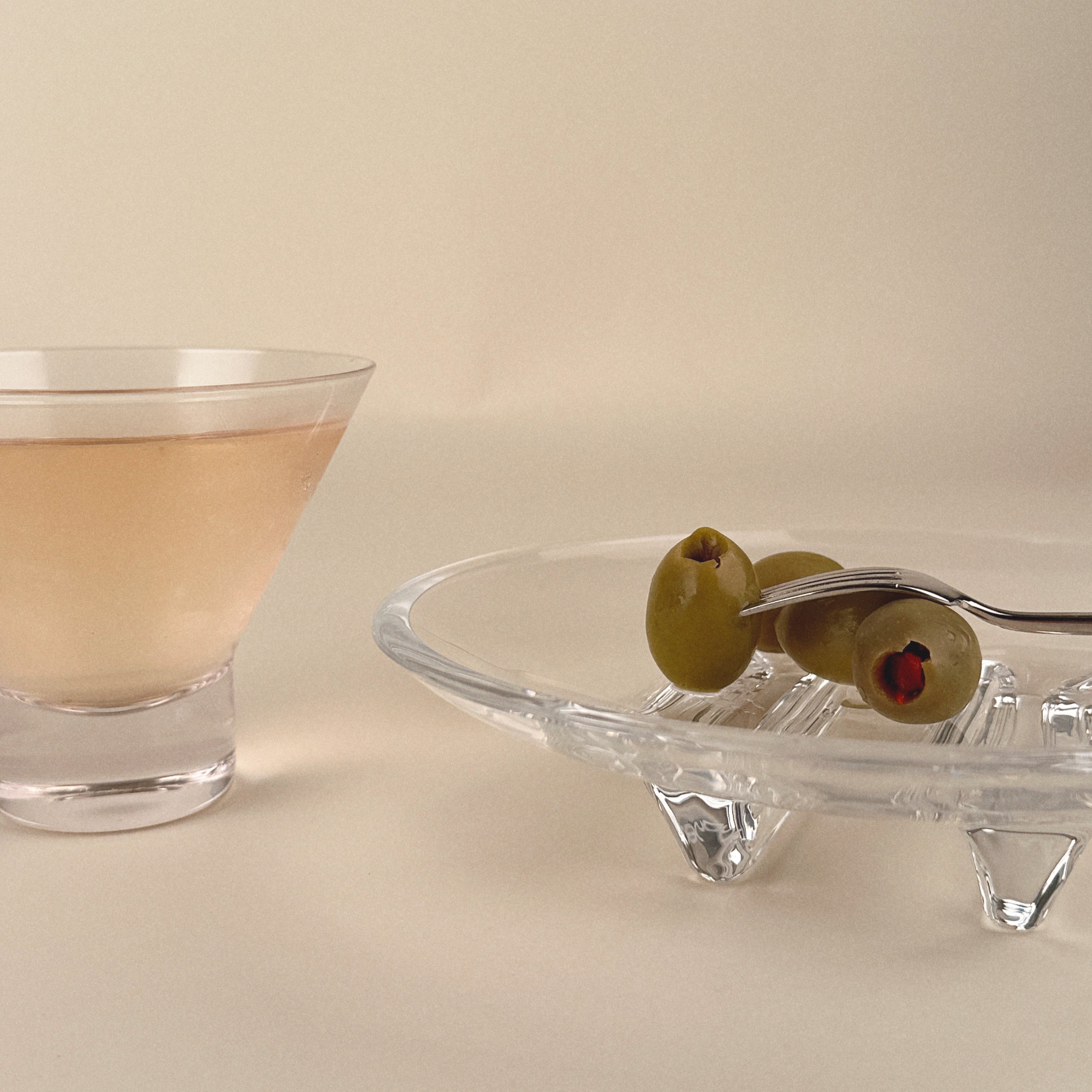 Cocktail glass with a drink and olives on a clear dish against a beige background