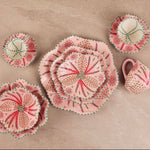A set of handmade fabric table linens, including plates, a napkin, and a small pitcher, featuring a pink and red floral pattern with green and white accents, arranged on a beige stone surface.