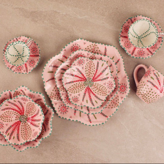 A set of handmade fabric table linens, including plates, a napkin, and a small pitcher, featuring a pink and red floral pattern with green and white accents, arranged on a beige stone surface.