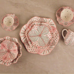 A set of handmade ceramic tableware with a distinctive hand-painted patchwork design in pink, red, and green on a beige background, arranged on a neutral stone surface.