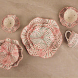 A set of handmade ceramic tableware with a distinctive hand-painted patchwork design in pink, red, and green on a beige background, arranged on a neutral stone surface.
