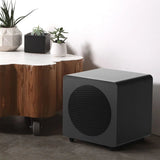 Black subwoofer next to a wooden coffee table with plants on a light-colored floor.