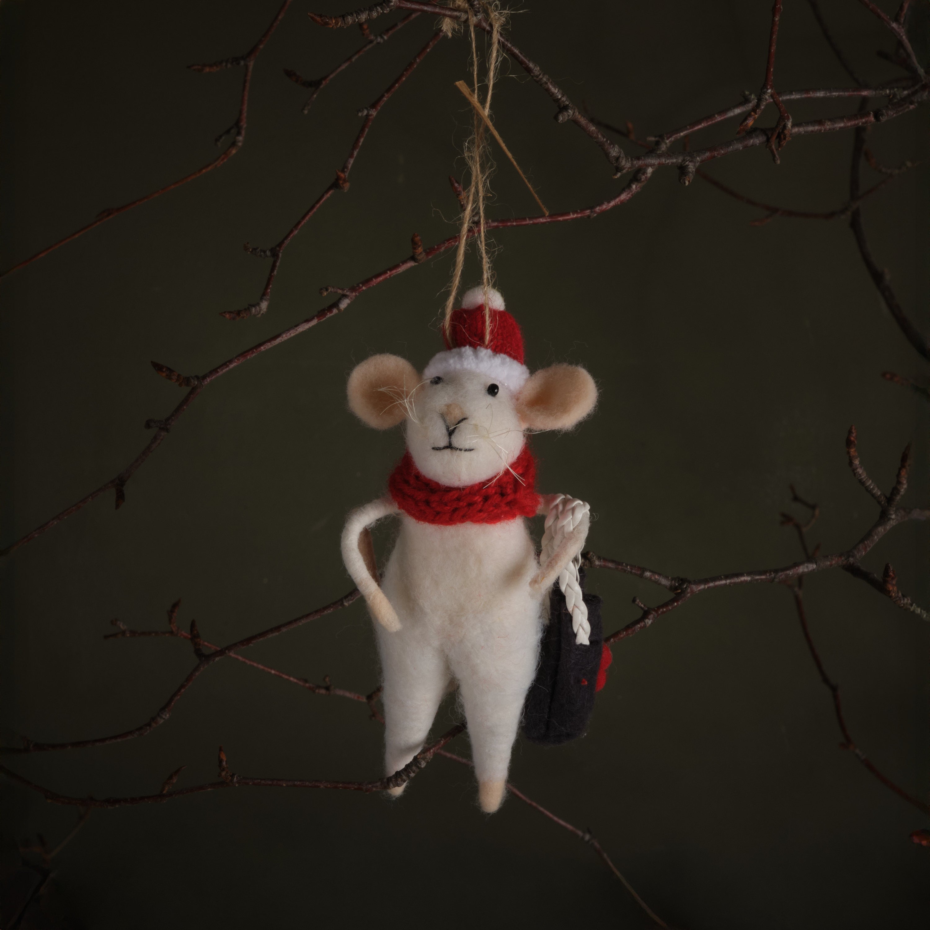 White mouse ornament with red hat and scarf hanging on a branch against a dark background