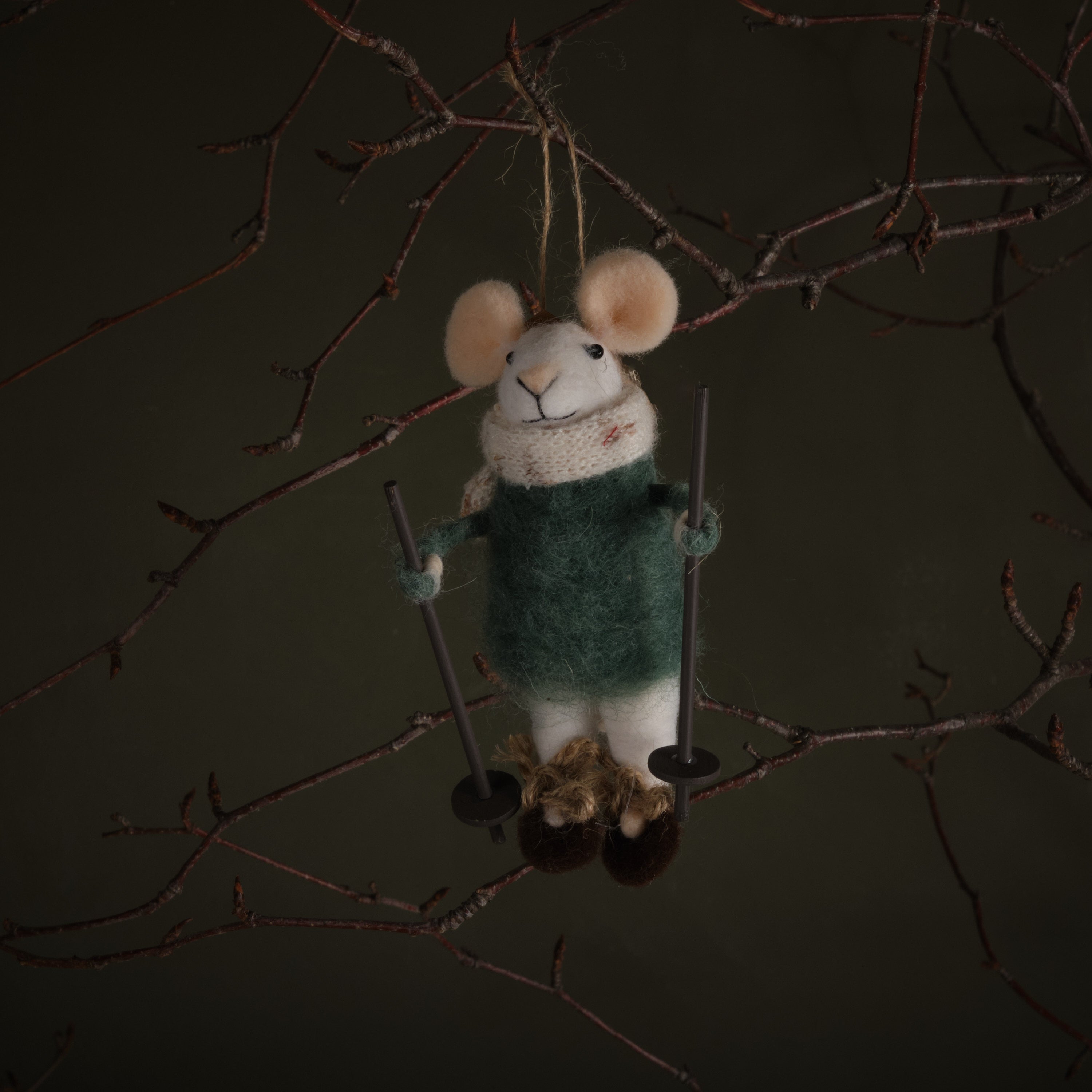 Felted mouse ornament in a green sweater hanging from a branch against a dark background