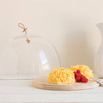 Yellow cake with raspberries under a glass cloche on a wooden board, next to a white ceramic pitcher.