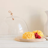 Yellow cake with raspberries under a glass cloche on a wooden board, next to a white ceramic pitcher.
