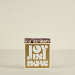 Matchbox with 'Joy is Now' text on a beige background