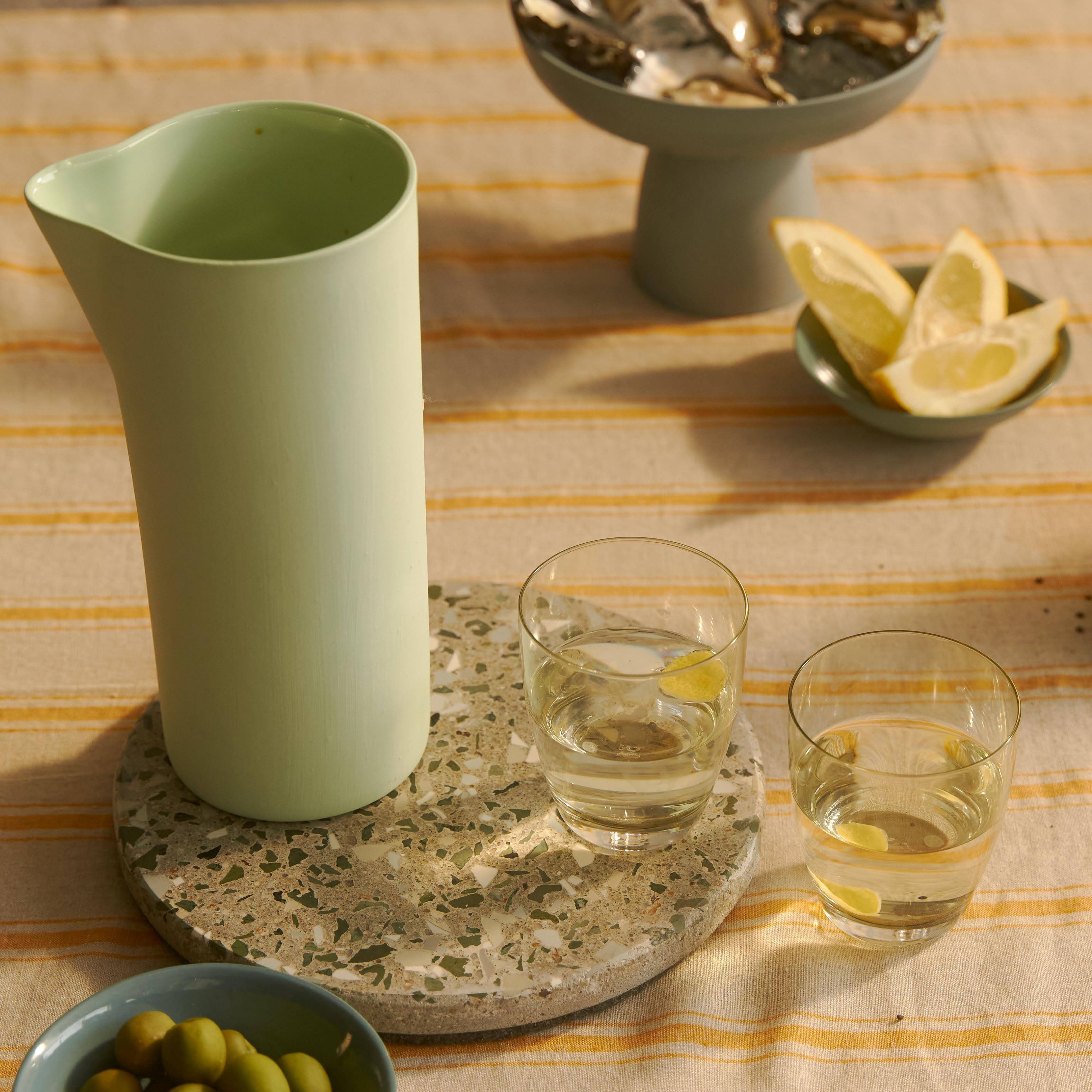 Green pitcher on a stone coaster with two glasses of liquid and bowls of lemons and olives on a striped tablecloth.