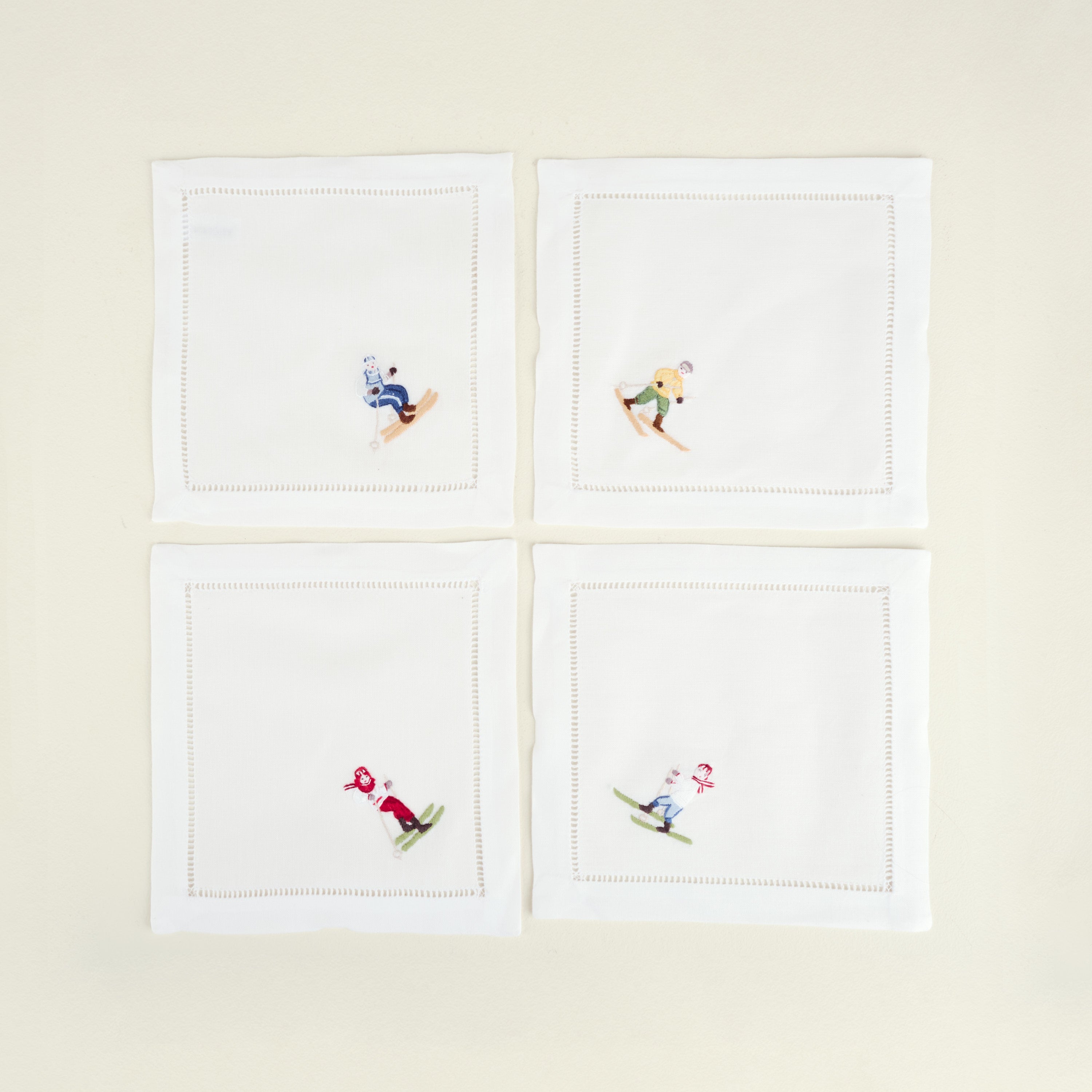 Four white napkins with embroidered skiing designs on a beige background