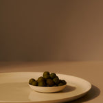 Small white bowl filled with green olives on a beige surface with a brown background