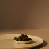 Small white bowl filled with green olives on a beige surface with a brown background