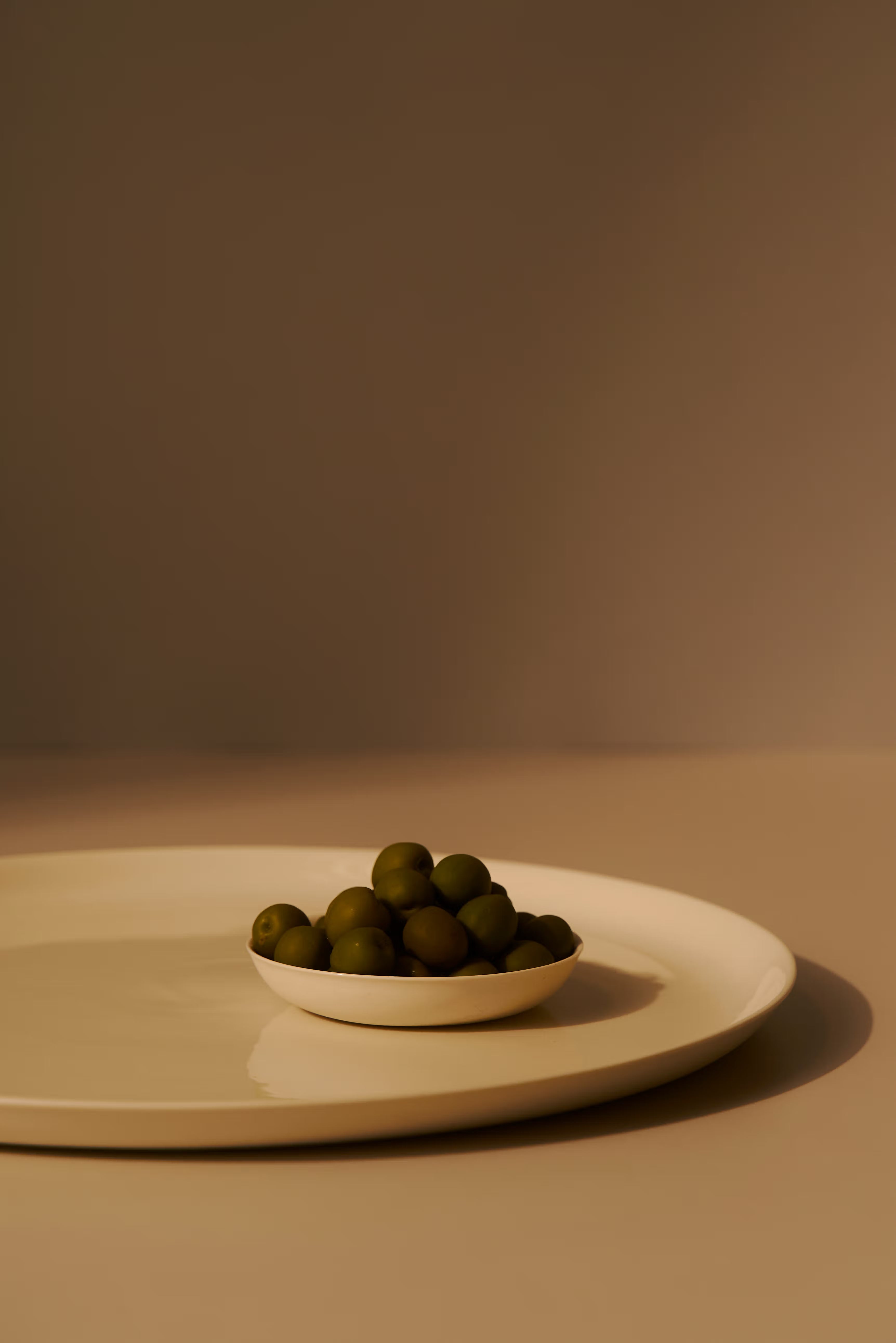 Small white bowl filled with green olives on a beige surface with a brown background