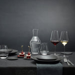 Dining table set with wine glasses, decanter, and black plates on a dark background