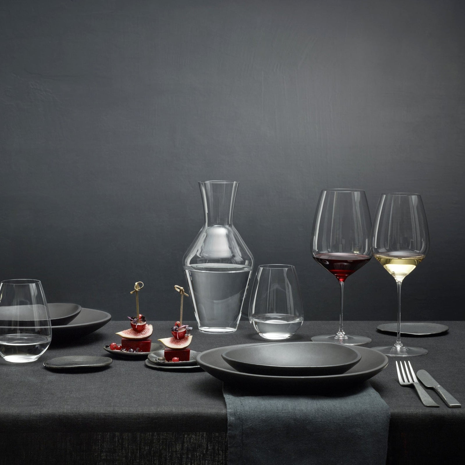 Dining table set with wine glasses, decanter, and black plates on a dark background