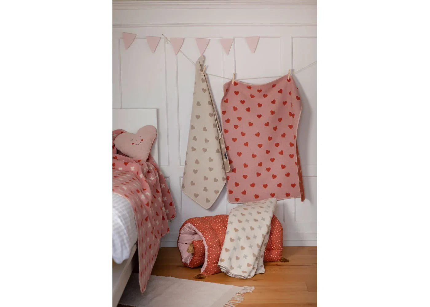 Children's blankets with heart patterns on a wooden floor against a white wall with pink flags.