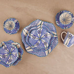 A set of handmade ceramic tableware with a blue and white geometric mosaic pattern, including a large plate, a small plate, a bowl, a cup, and a small dish, arranged on a beige stone tile background.