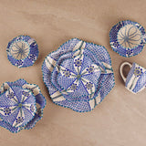 A set of handmade ceramic tableware with a blue and white geometric mosaic pattern, including a large plate, a small plate, a bowl, a cup, and a small dish, arranged on a beige stone tile background.
