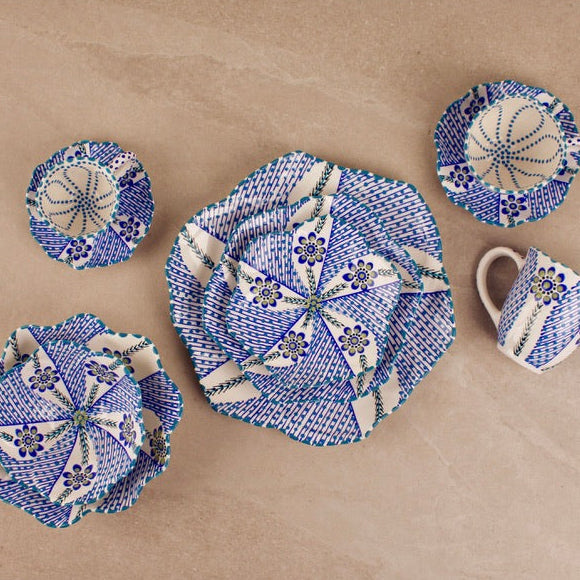 A set of handmade ceramic tableware with a blue and white geometric mosaic pattern, including a large plate, a small plate, a bowl, a cup, and a small dish, arranged on a beige stone tile background.
