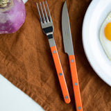 Orange-handled fork and knife set on a brown cloth with a purple turnip and white plate in the background.