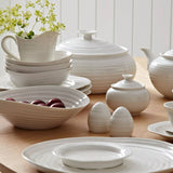 Set of white ceramic tableware including bowls, plates, and teapots on a wooden surface.