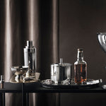 Modern bar setup with cocktail shaker, bottles, and glassware on a dark surface.