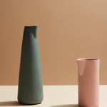 Two ceramic vases, one green and one pink, on a beige surface with a brown background.