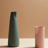 Two ceramic vases, one green and one pink, on a beige surface with a brown background.