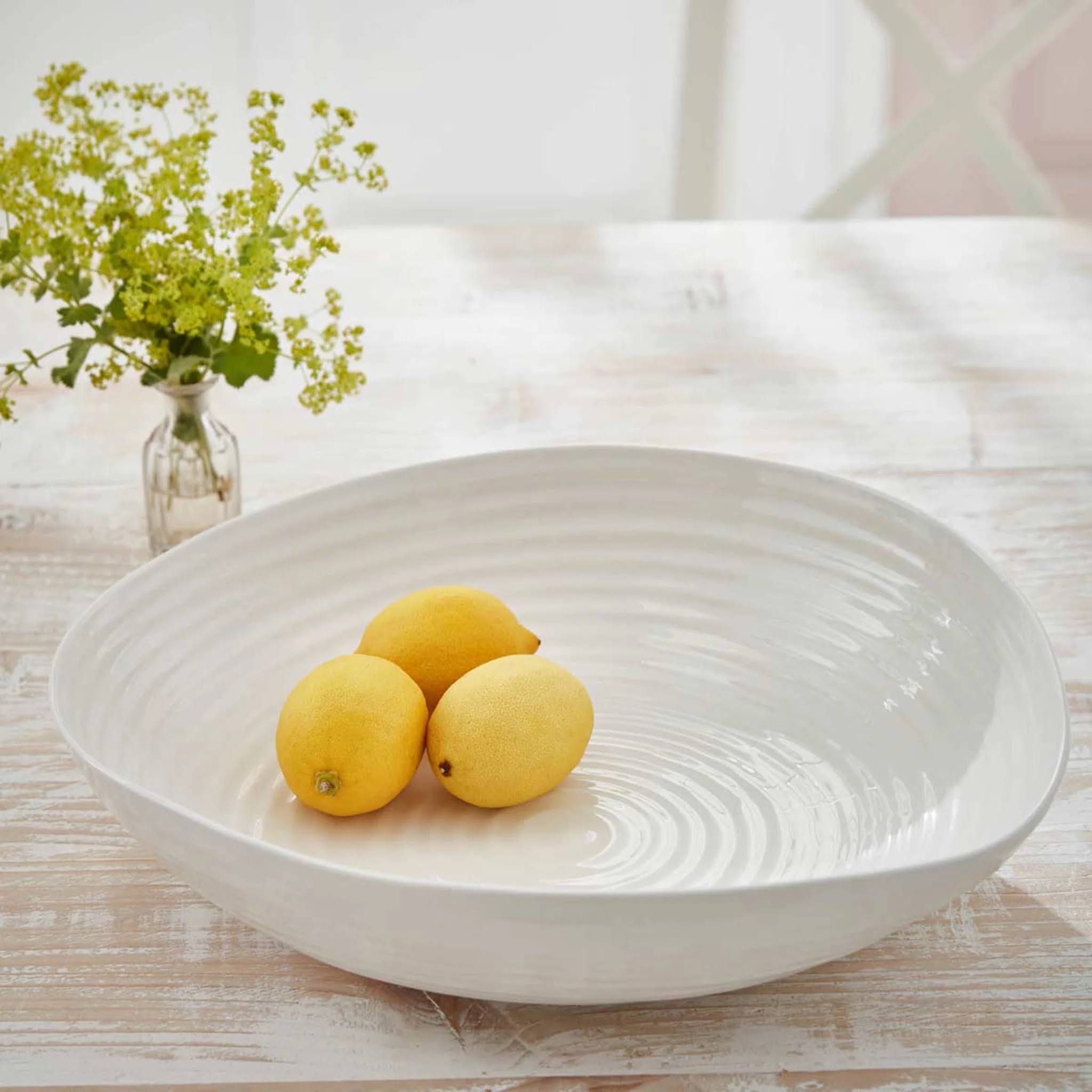 White ceramic bowl with lemons on a wooden table, next to a vase with greenery.