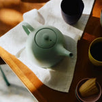 Green teapot on a white cloth with a wooden table and other items in the background
