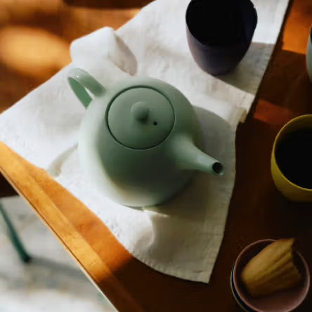Green teapot on a white cloth with a wooden table and other items in the background