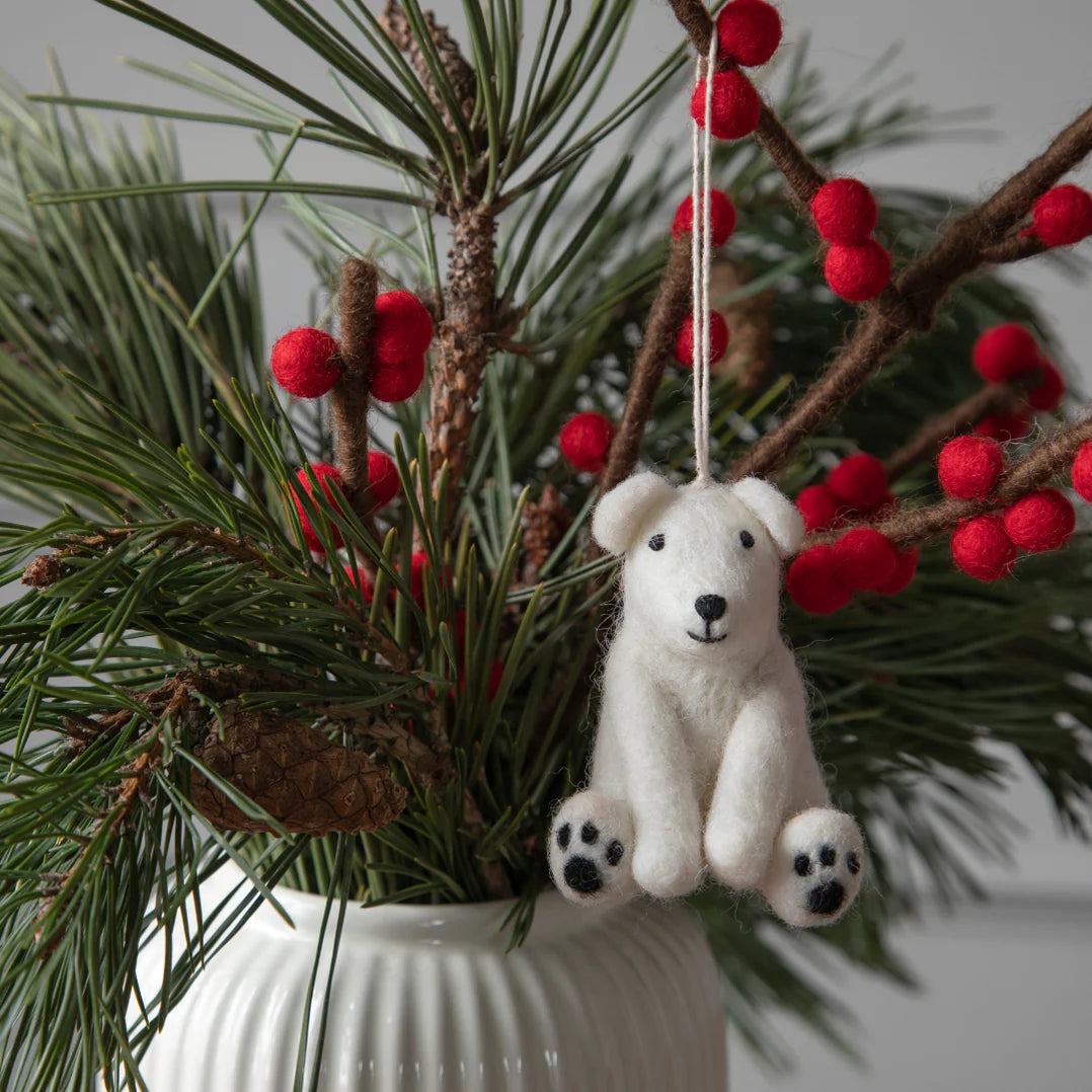A white felt polar bear ornament hanging from a thread, displayed amidst green pine needles and red berries in a white vase.